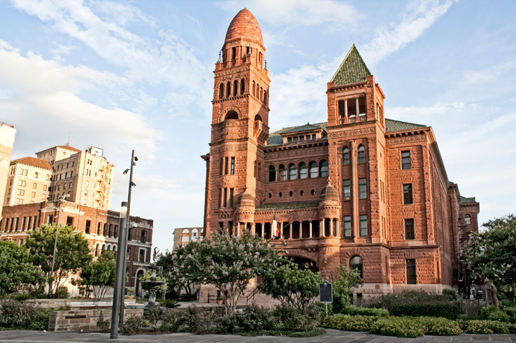 A setting sun casts an orange glow on the old Bexar County Courthouse, a Romanesque-style building made of red sandstone.