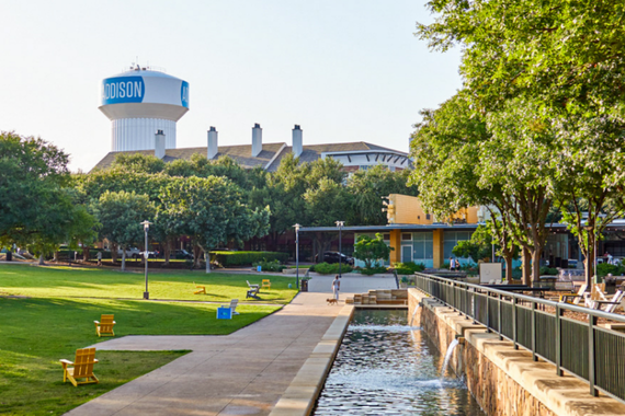 Water fountains stretch through Addison Circle Park, framed by manicured lawns and vibrant pink flower beds on a sunny day.