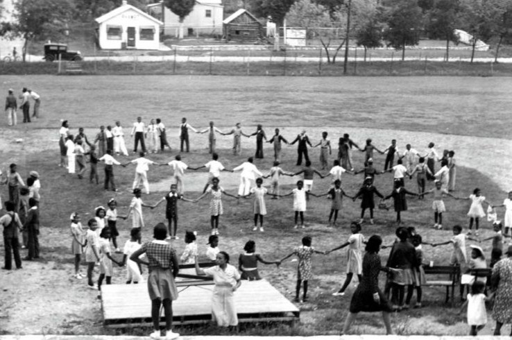 A black-and-white archival photo of children holding hands in concentric circles during a community gathering at Rosewood Neighborhood Park in East Austin, with the historic Henry G. Madison Cabin visible in the background.