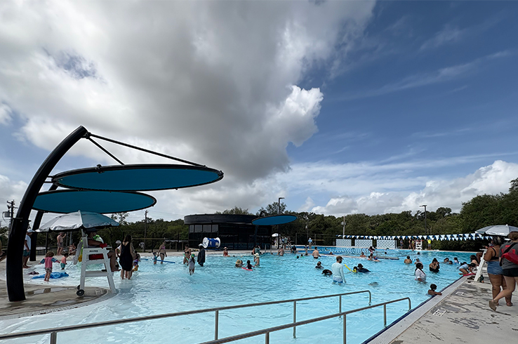 Swimmers fill the refurbished outdoor pool at Givens District Park in East Austin under modern blue shade canopies on a partly cloudy day.