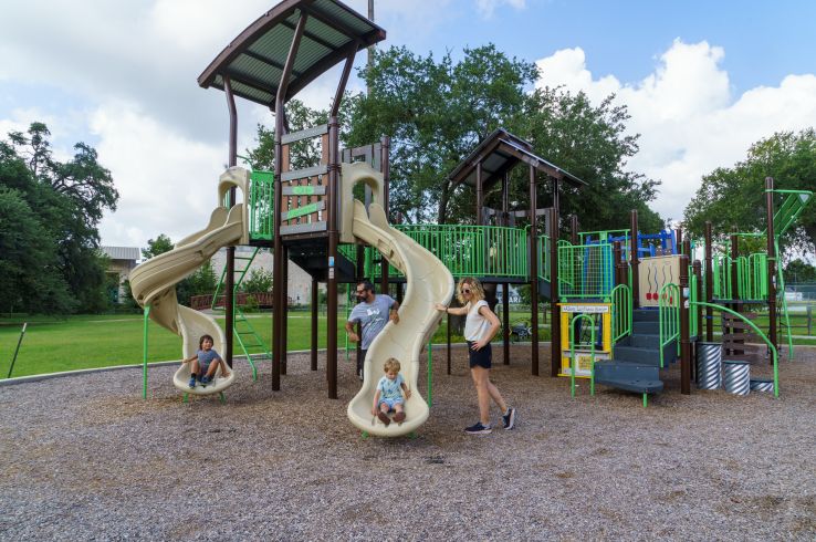 A family plays at the modern playground at Parque Zaragoza in East Austin, with children on tan twisting slides and a parent looking on.