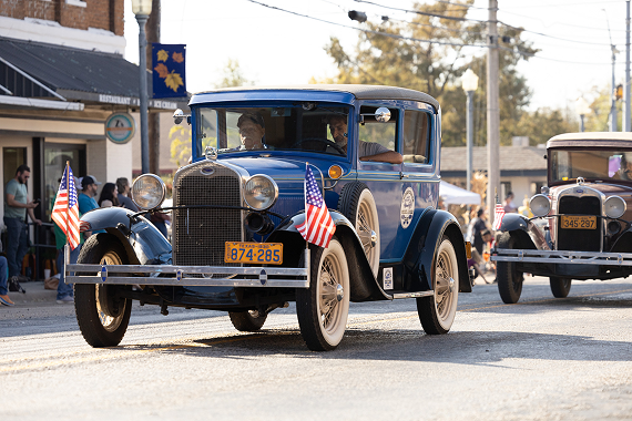 A blue antique Ford leads the antique car parade down Main Street during Winnsboro Autumn Trails, with a second vintage car following and spectators lining the sidewalk.