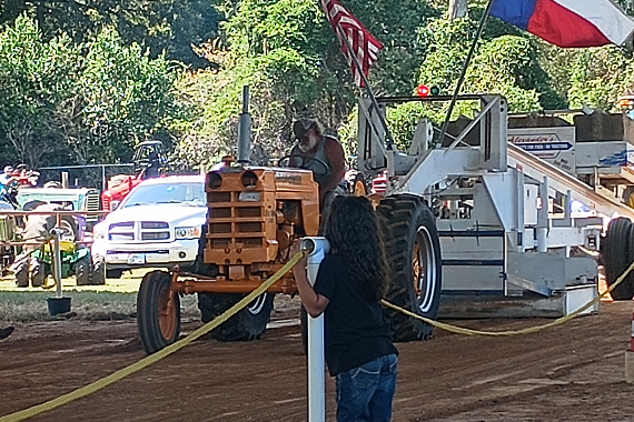 A vintage orange tractor competes in a tractor pull during Winnsboro Autumn Trails, with American and Texas flags flying and spectators watching from behind a rope line.