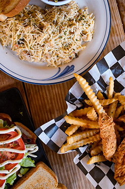 An overhead spread of dishes at a local Alvin restaurant, including a cheese-topped pasta, crinkle-cut fries with fried chicken tenders, and a caprese-style sandwich on a wooden table.