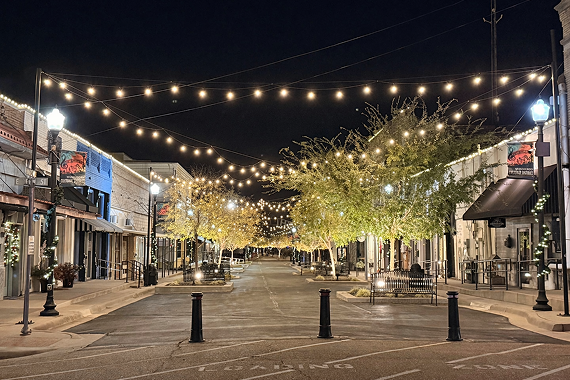 String lights hang over a brick-paved street in Winnsboro's Cultural Arts District at night, with lit trees, benches, and storefronts lining both sides.