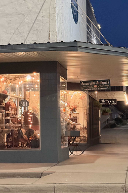 The lit corner storefront of Amaryllis Antiques at dusk in downtown Winnsboro, its window display glowing with string lights and a caf&eacute; sign visible next door.