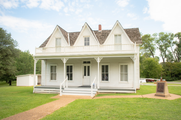 The Eisenhower Birthplace State Historic Site in Denison, Texas — a white two-story frame house with a covered front porch, three peaked dormers, and a historical marker on the manicured grounds where President Dwight D. Eisenhower was born in 1890.