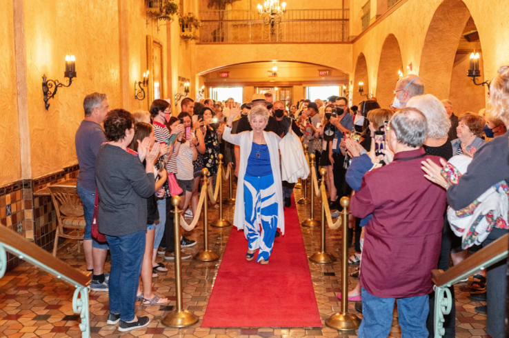 A guest walks the red carpet through the Plaza Theatre lobby as attendees applaud from behind velvet ropes, with the theatre's warm Spanish Colonial arches, wrought-iron sconces, and mosaic tile floors visible in the background.