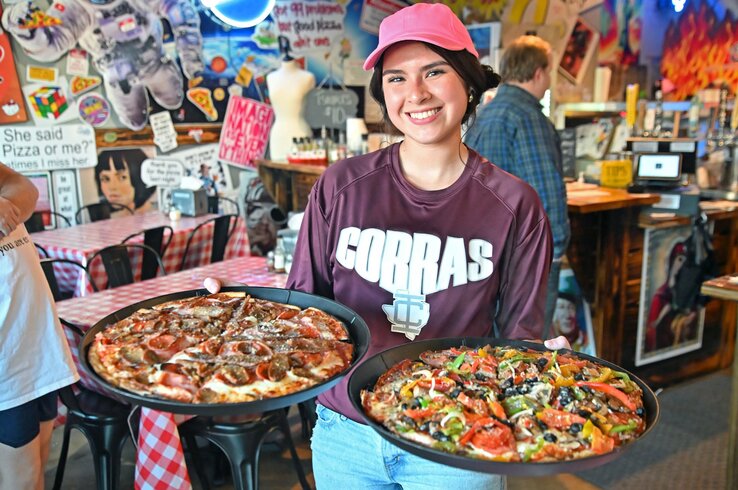 A smiling server carries two large pizzas — one meat-topped and one loaded with vegetables — through a vibrant, pop-art-decorated pizza restaurant with checkered tablecloths.