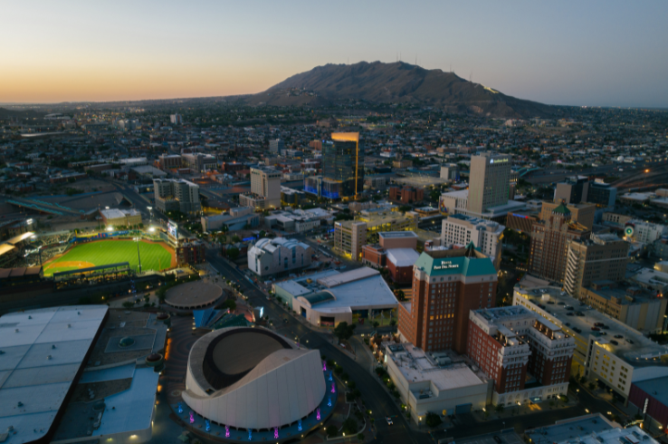 Aerial view of downtown El Paso, Texas at dusk, with the city skyline, Southwest University Park baseball stadium, and the Franklin Mountains rising in the background.