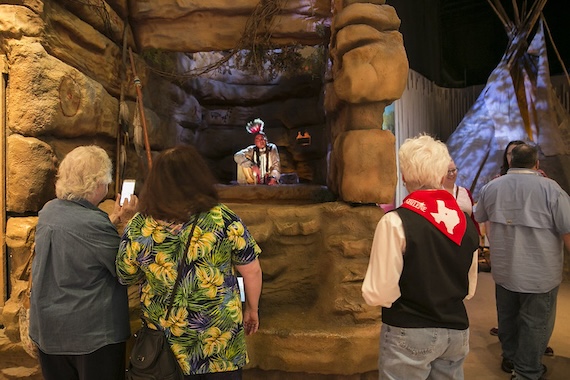 Museum visitors gather at a rock-formation exhibit at Frontier Texas to watch a holographic Spirit Guide in Native American dress, with a tipi visible in the gallery beyond.