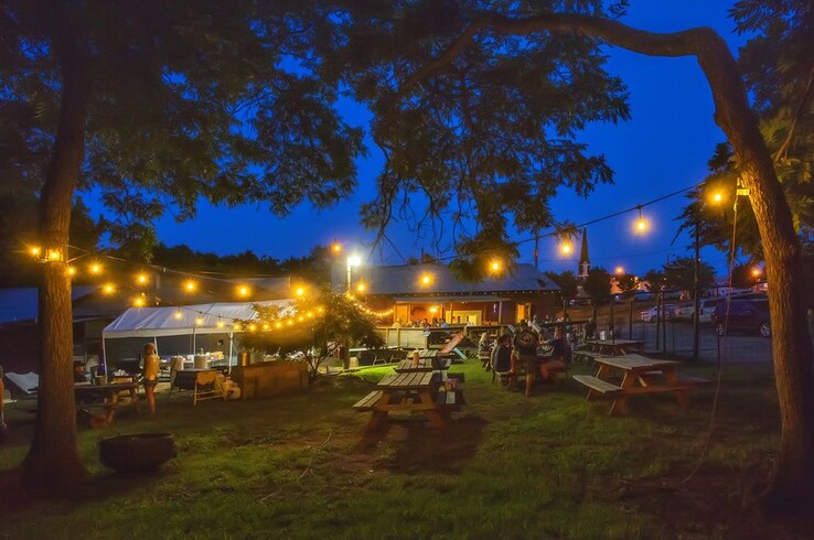 Hambones restaurant in Old Town Palestine, Texas, at dusk with picnic tables, string lights strung between mature trees, and a covered bar area glowing in the background.