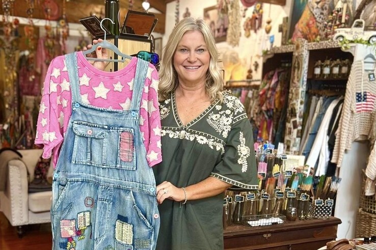 A boutique shop owner holds up a pair of patchwork denim overalls over a pink star-print top inside a colorful retail store filled with clothing, accessories, and home goods.