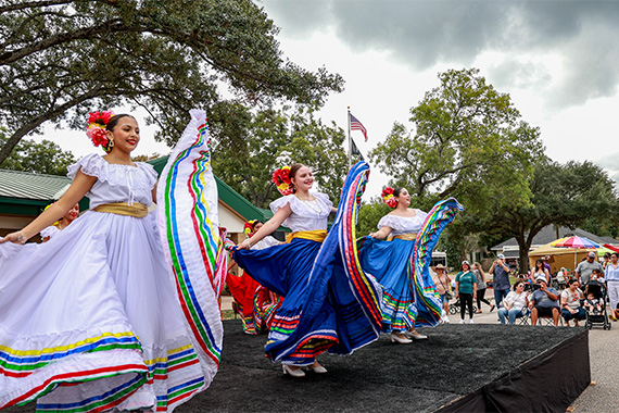 Ballet Folklórico dancers in flowing white and blue dresses with colorful ribbon trim perform on an outdoor stage at Alvin's Hispanic Heritage Fiesta, with spectators watching from the sidewalk.