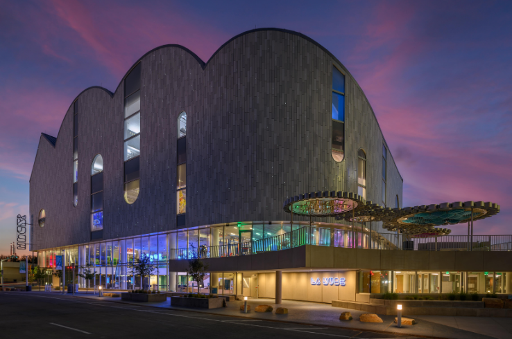 The cloud-shaped exterior of La Nube STEAM Discovery Center lit at dusk in downtown El Paso, with a scalloped roofline, glowing ground-floor windows, and a colorful canopy sculpture at the entrance.