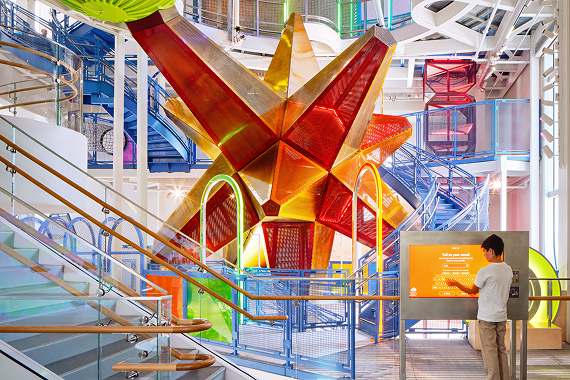 The Anything's Possible Climber at La Nube, a multi-story structure of brightly colored geometric forms and mesh panels, with a visitor using a touchscreen exhibit in the foreground.