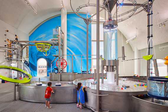 Two children play at a large stainless steel water table in the Flow gallery at La Nube, surrounded by pumps, pipes, and a vertical water column against a blue mural wall.