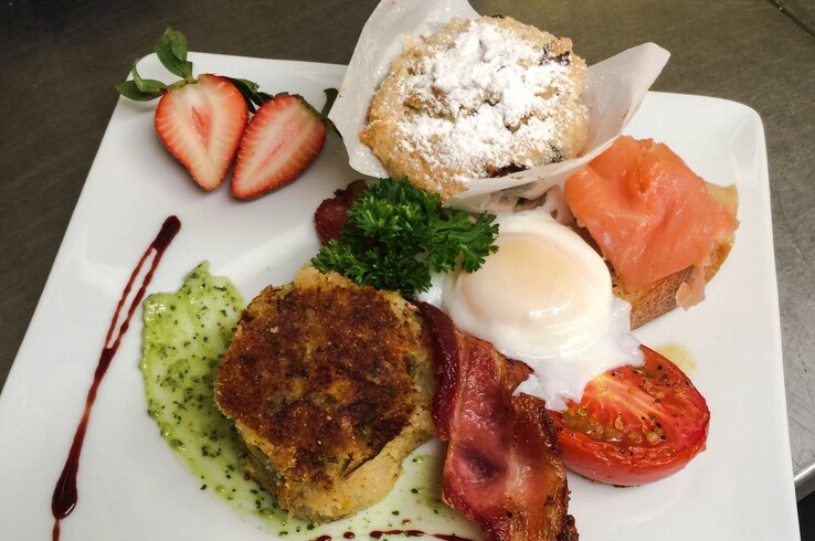 Plated dish at Restaurant Aubergine at Sabor a Pasión in Palestine, Texas, featuring a golden-brown crab cake, poached egg, crispy bacon, smoked salmon on toast, roasted tomato, powdered sugar muffin, and sliced strawberry with herb purée and balsamic drizzle.