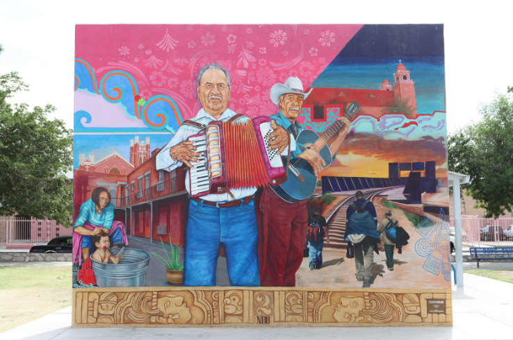 A large mural in El Paso's Segundo Barrio depicting two musicians with an accordion and guitar, surrounded by scenes of neighborhood life, a church, and railroad tracks, with a carved Mesoamerican-style base.