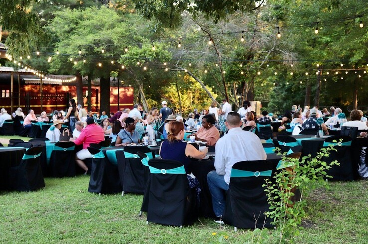 Outdoor evening dinner event at the Texas State Railroad in Palestine, Texas, with guests seated at round tables draped in black linens with teal sashes under string lights and a tree canopy near the depot.