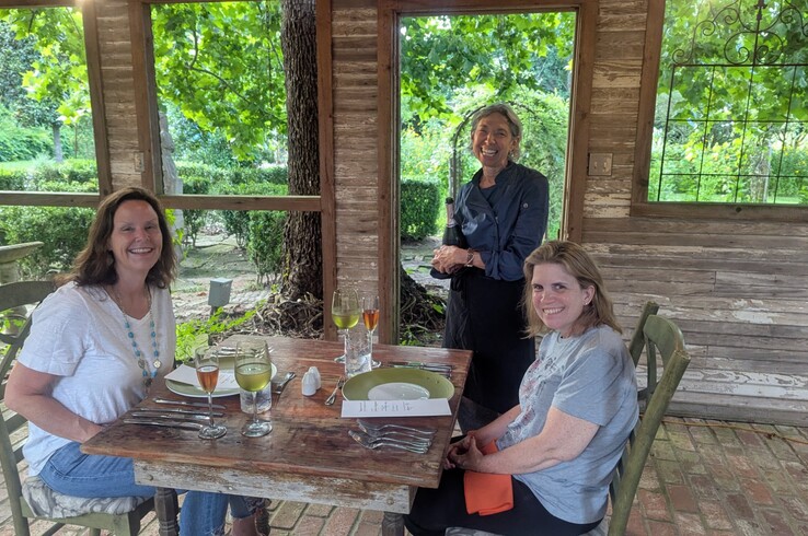 Guests seated at a rustic wooden table at Texas Foodie Adventures at Hummingbird Hollow in Palestine, Texas, with wine glasses and place settings as a server presents a bottle in the shaded outdoor dining pavilion surrounded by gardens.