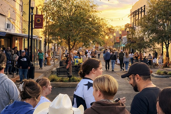 Crowds fill the Bowery in downtown Winnsboro at sunset, with banners on the lampposts, string lights overhead, and shops open along both sides of the street.